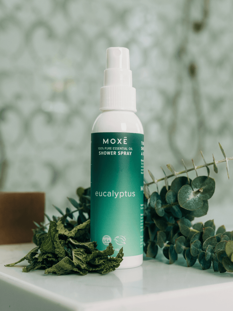 MOXĒ Eucalyptus Aromatherapy Shower Spray in front of fresh eucalyptus and mint leaves