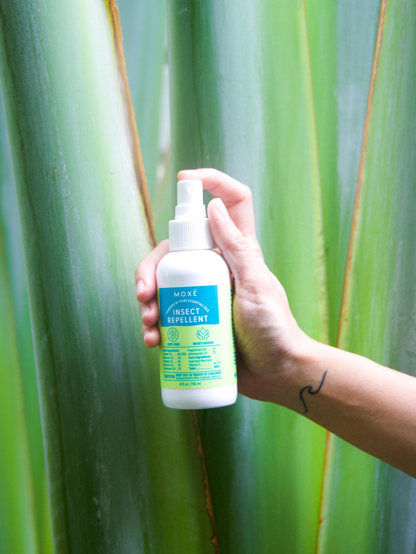 Hand holding MOXĒ Insect Repellent in front of palm leaves